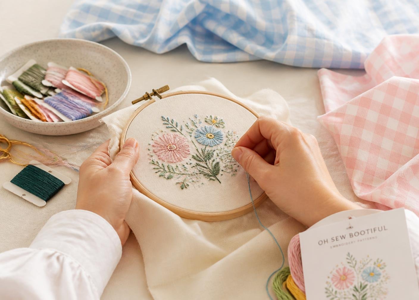 Hands embroidering pastel flowers on cream fabric beside gingham cloth